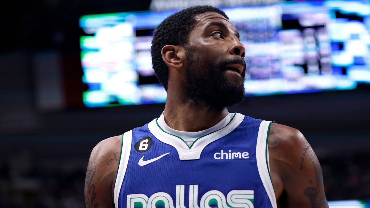 A Black man in a blue and white Dallas Mavericks jersey looks away from the camera on an NBA basketball court.