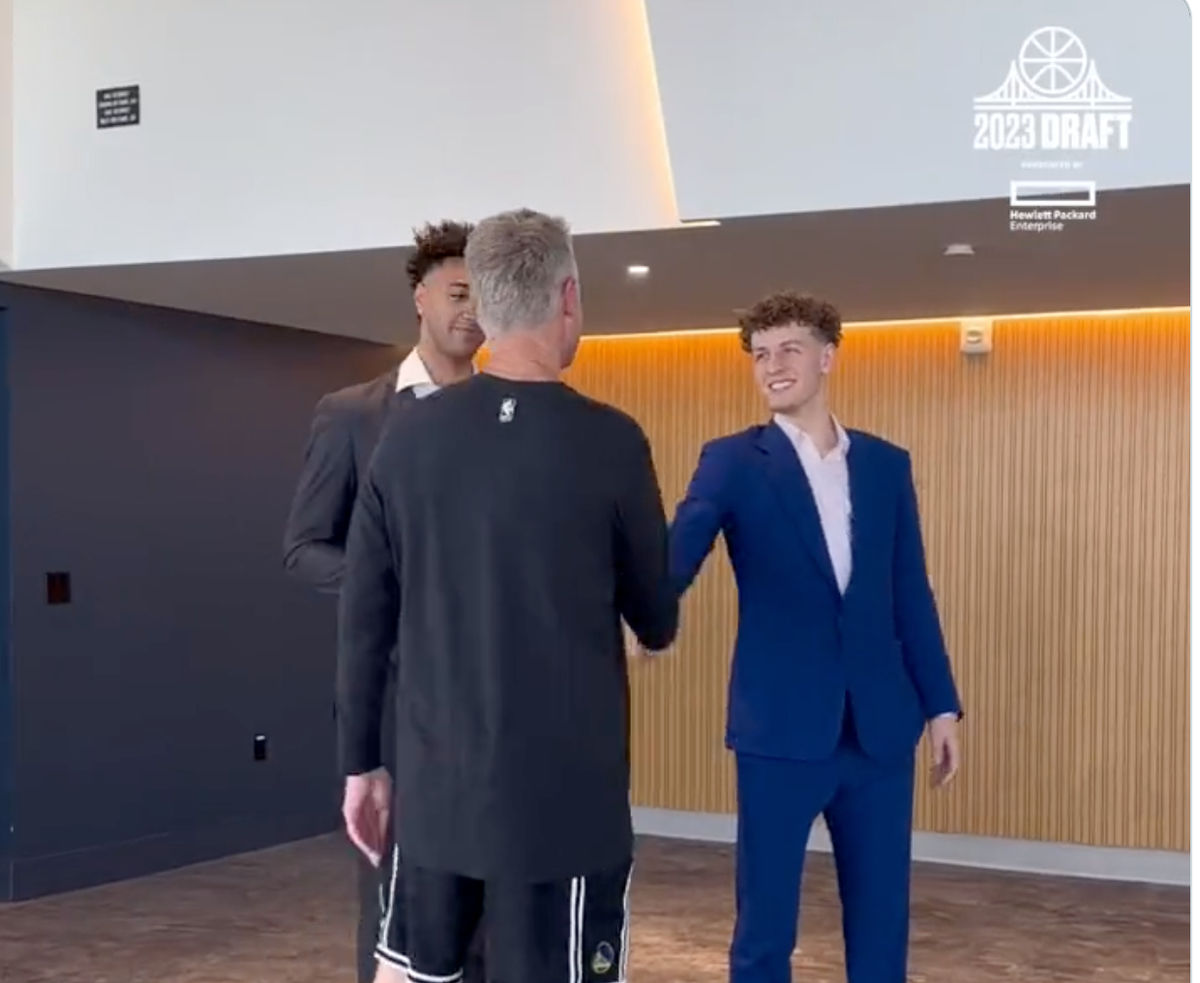 A white man, dressed in a black long-sleeved t-shirt and black basketball shorts, shakes the hand of a young man in a blue suit.
