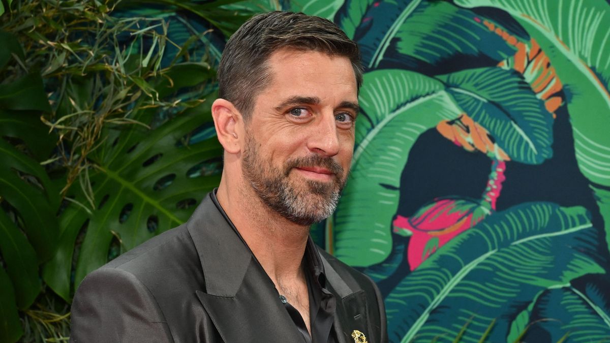A white man with brown hair and a shiny black suit smirks at the camera in front of a backdrop of leafy greens.