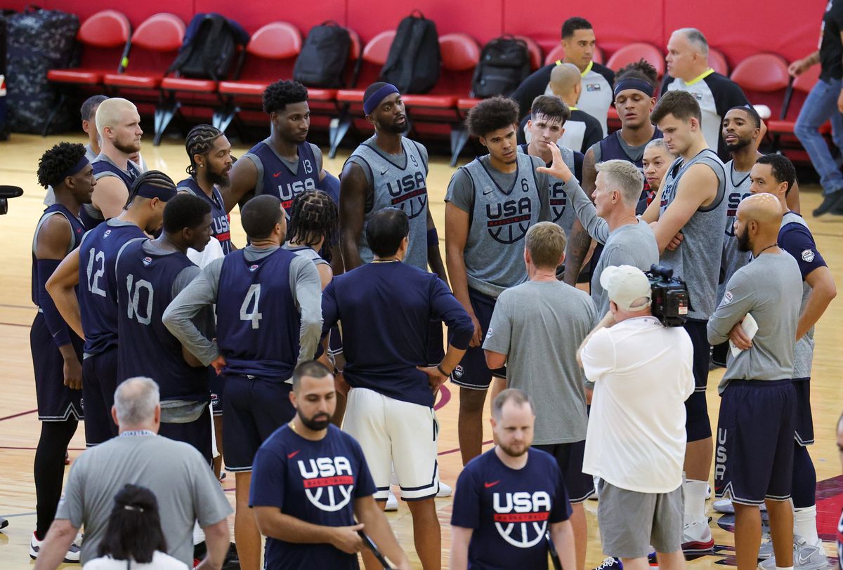 A group of men in navy blue jerseys and black basketball shorts stand in a huddle on a basketball court and listen to a man gesticulating in the center of a group.