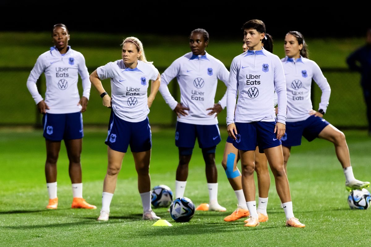 Six women of different heights and races, wearing matching gray pullovers with blue shorts, warm up with soccer balls at their feet.