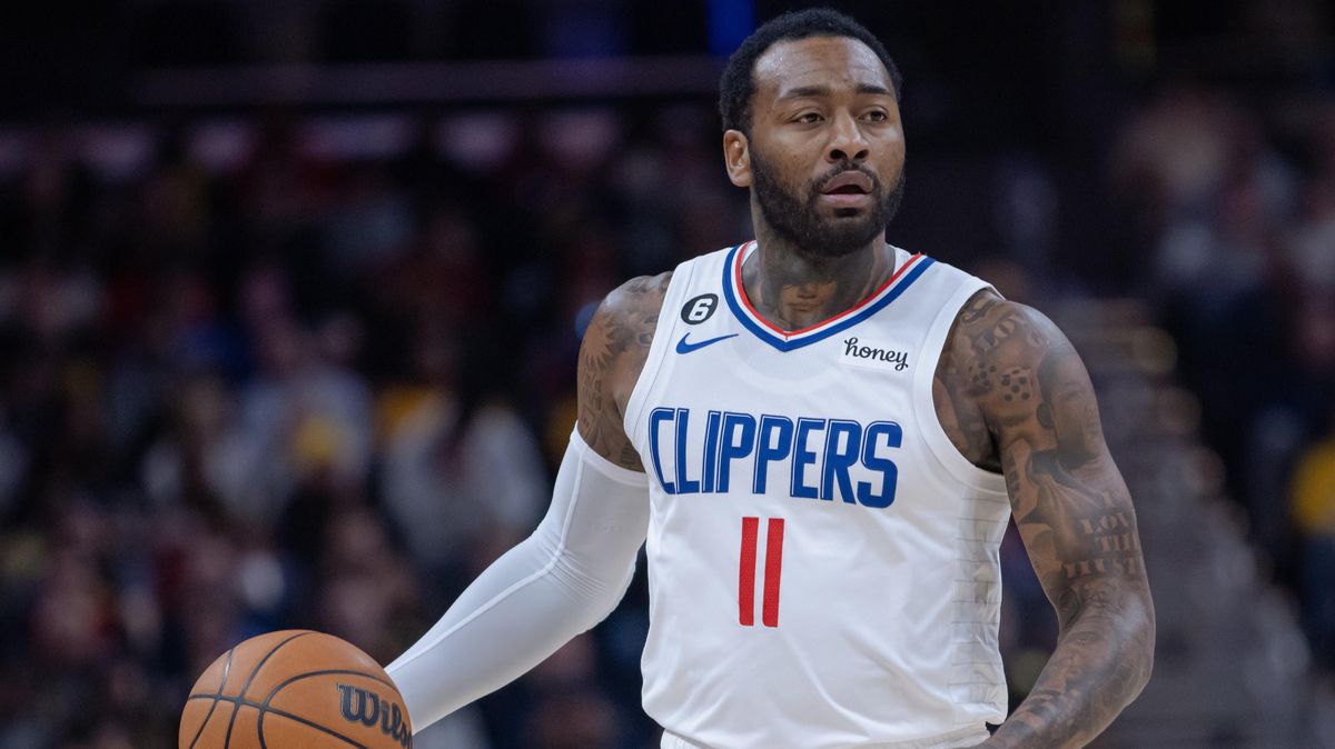 A Black man in a white LA Clippers jersey with blue letters and red numbers dribbles a basketball at center court.