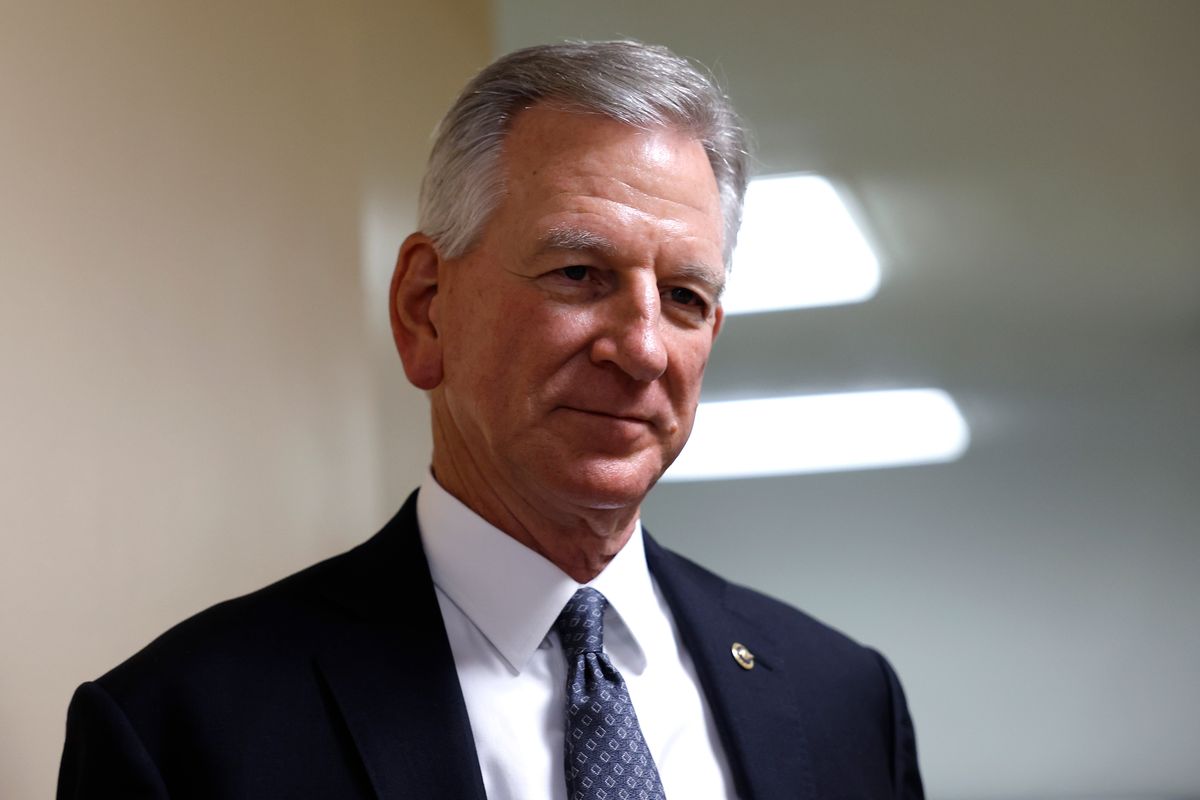 A white man with gray hair in a black suit with a blue tie stands with a dumb look on his face.