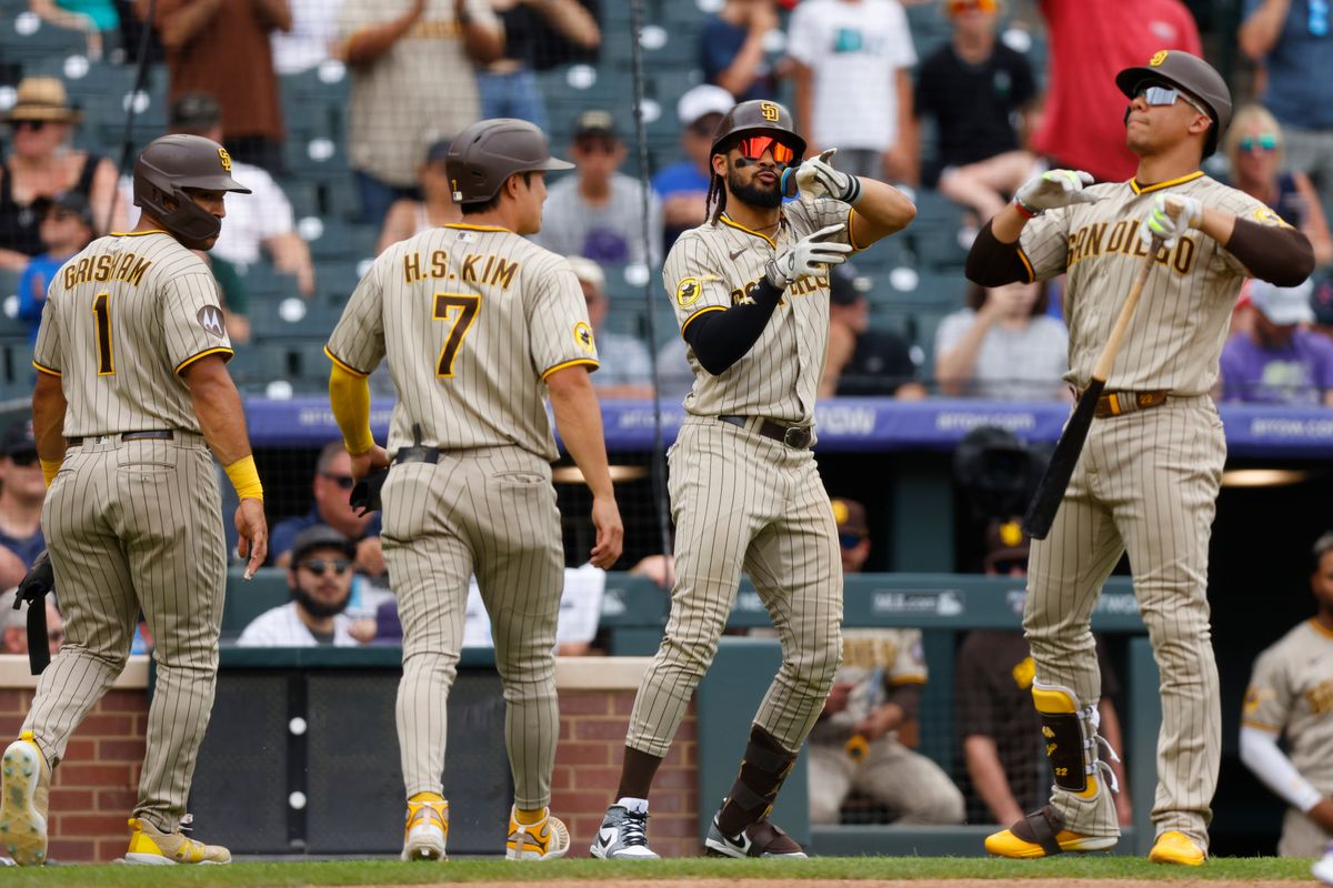 Two baseball players, in gray uniforms with brown stripes and helmets, walk back to the dugout while two of their teammates make celebratory gestures beside them.