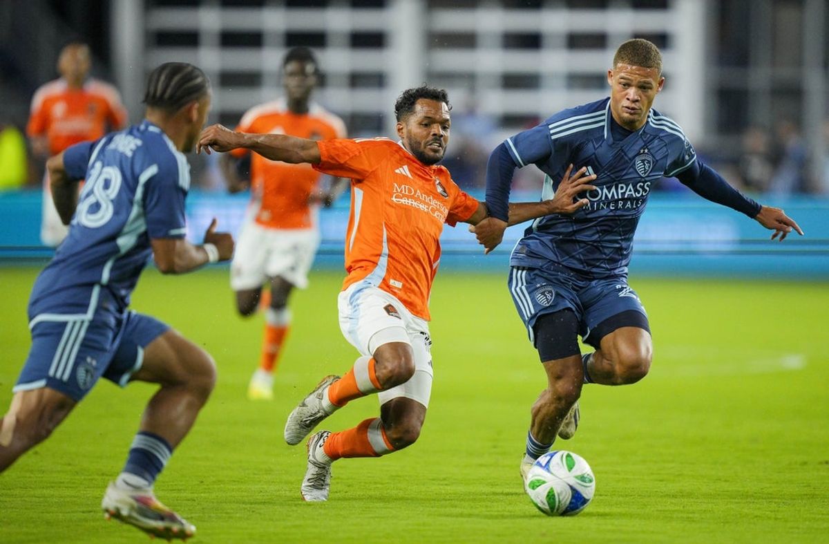 Oct 18, 2025; Kansas City, Kansas, USA; Houston Dynamo midfielder Duane Holmes (14) fights for the ball against Sporting Kansas City defender Logan Ndenbe (18) and defender Zorhan Bassong (22) during the first half at Children's Mercy Park. Mandatory Credit: Jay Biggerstaff-Imagn Images