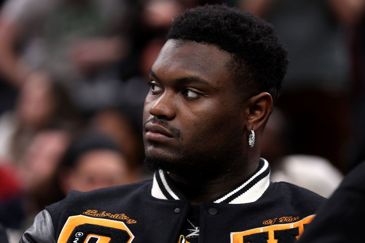 A young Black man with a diamond hoop earring and a black letterman jacket sits on the sidelines of an NBA game.