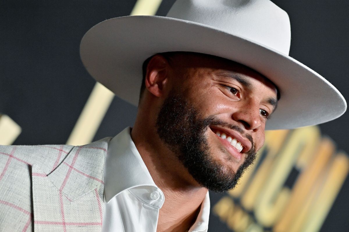 A Black man in a white cowboy hat and white shirt with pink window pan print smiles at at country music event.
