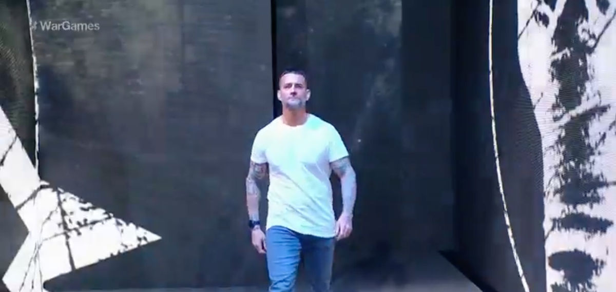 A man in a white t-shirt, jeans, brown hair and a salt-and-pepper close-cropped beard walks out before a large crowd against a black background at WWE's Survivor Series show in Chicago..