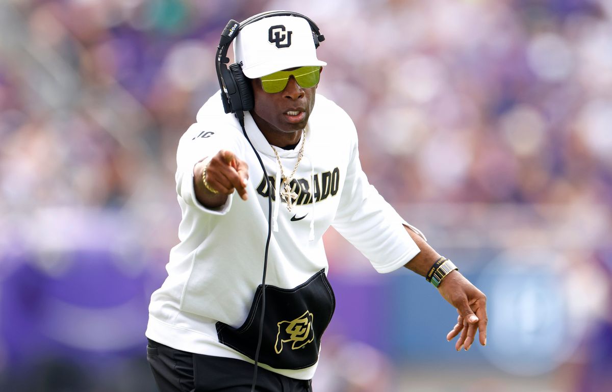 Colorado college football coach Deion Sanders, wearing sunglasses, a head set, and a white sweatshirt with black and gold detailing, points to his team from the sidelines of the Colorado-TCU game.