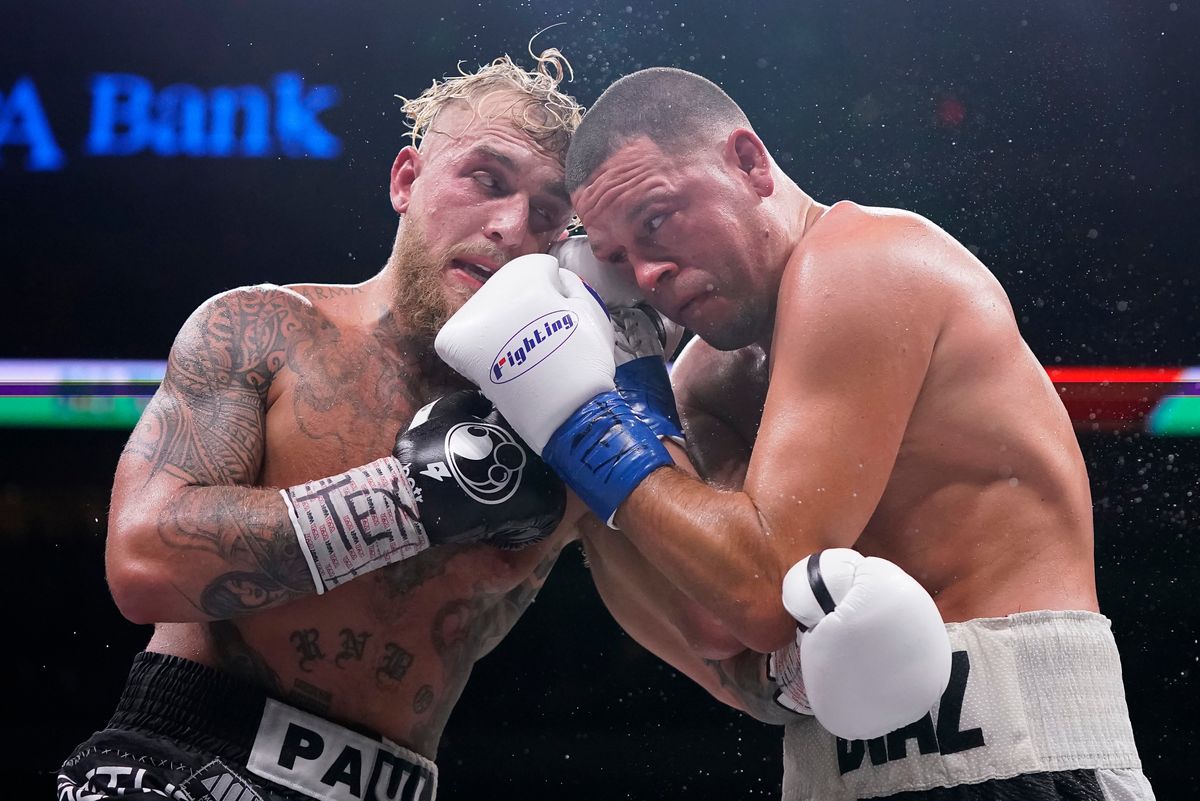 Two men, one blonde, one brunette, in boxing shorts and gloves lean into each other insider a boxing ring.