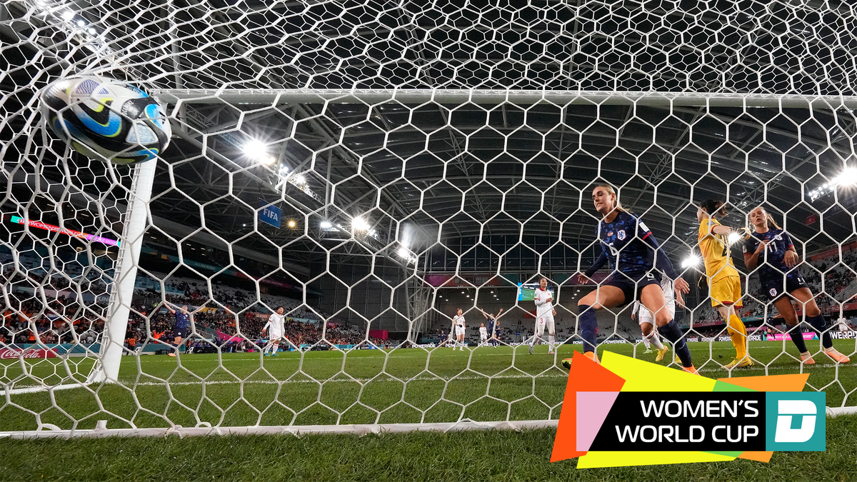 In a view from behind a soccer goal, a soccer ball looks as though it will burst through the net, as two women in matching blue soccer uniforms look on.