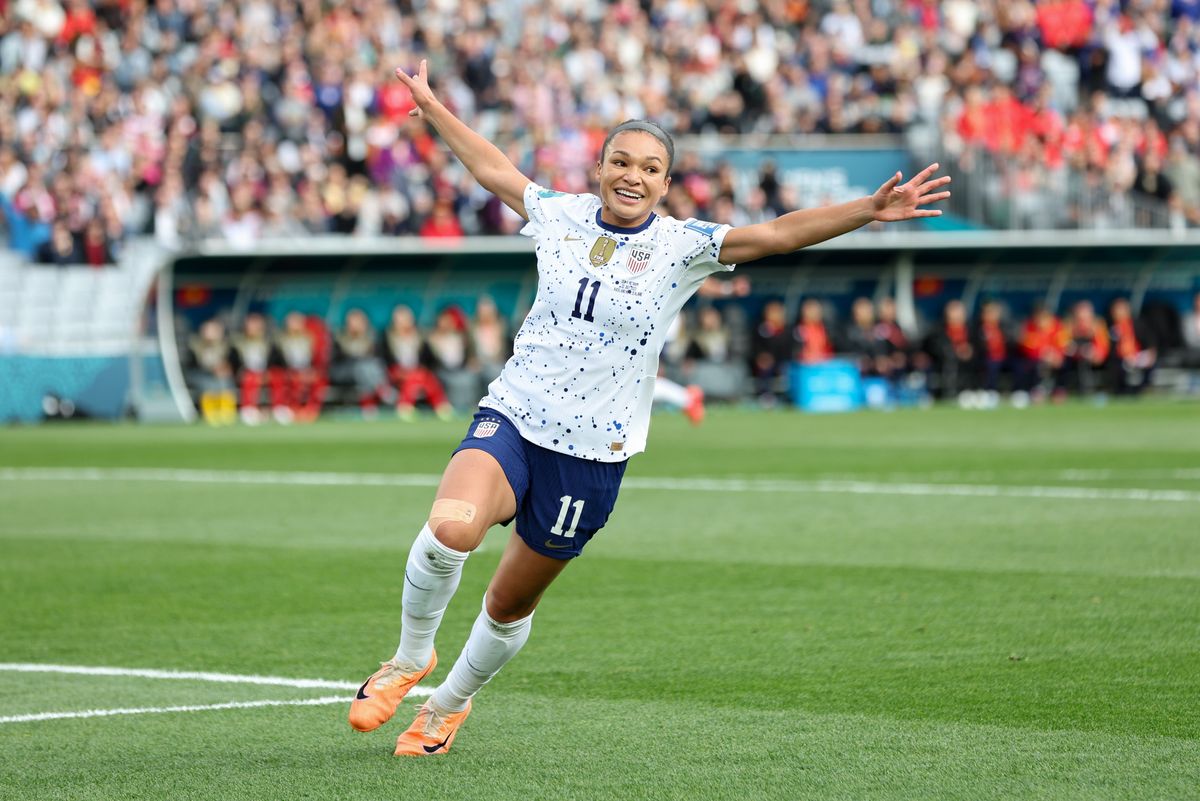 A young woman with dark hair pulled back in a braid, a white jersey with blue paint splatters and blue shorts holds her arms out wide in celebration on a soccer pitch.