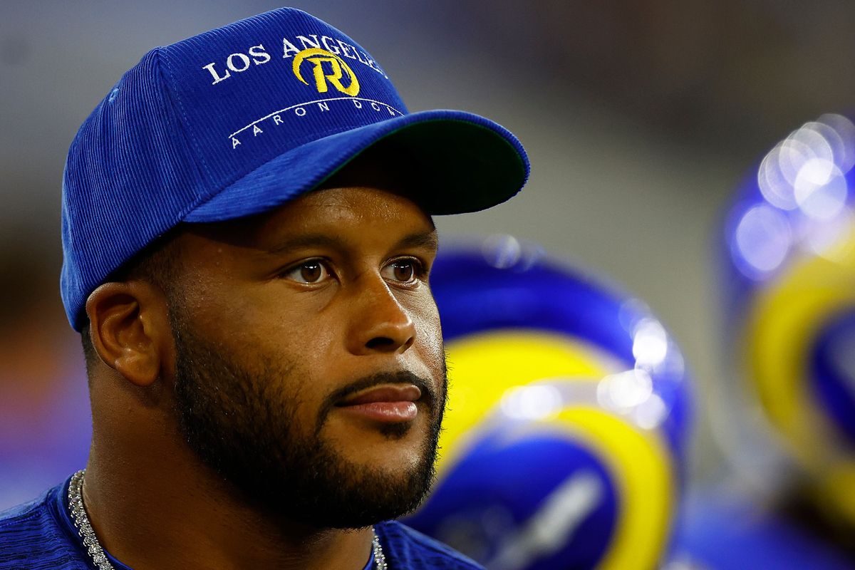 A Black man in a blue Los Angeles Rams baseball cap and a silver chain sits in front of blue and yellow Rams football helmets