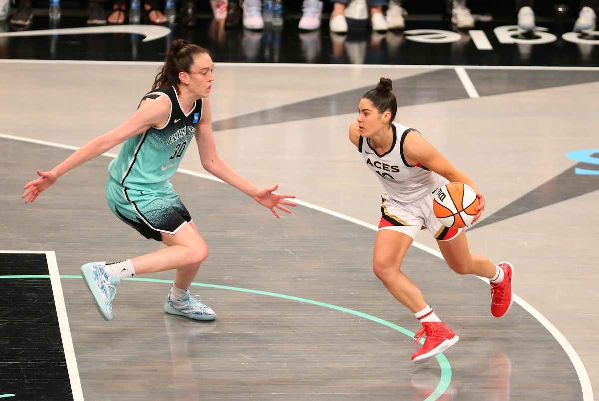 A white woman with a brown ponytail in a green and black uniform back peddles on a  basketball court, while a white woman with black hair in a ponytail and a white and black uniform dribbles a basketball towards her.