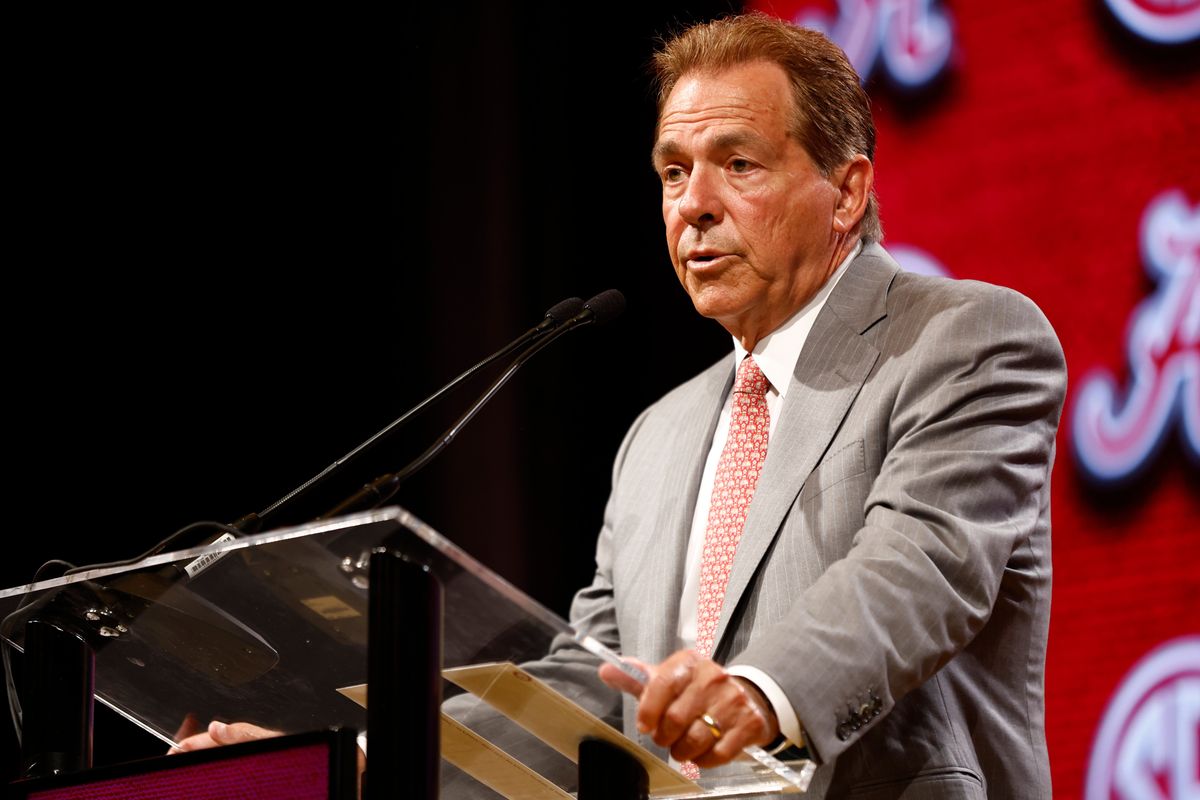 A white man in a gray suit and red patterned tie stands at a lectern in front of a red background.