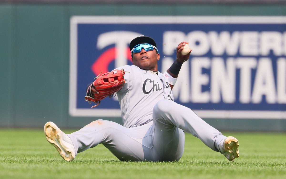 A Black man in a gray baseball uniform makes a throw while sitting on the ground on a baseball field.