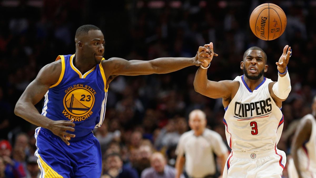 A Black man in a blue Warriors uniform knocks the basketball away from a Black man in a white Clippers uniform during an NBA basketball game.