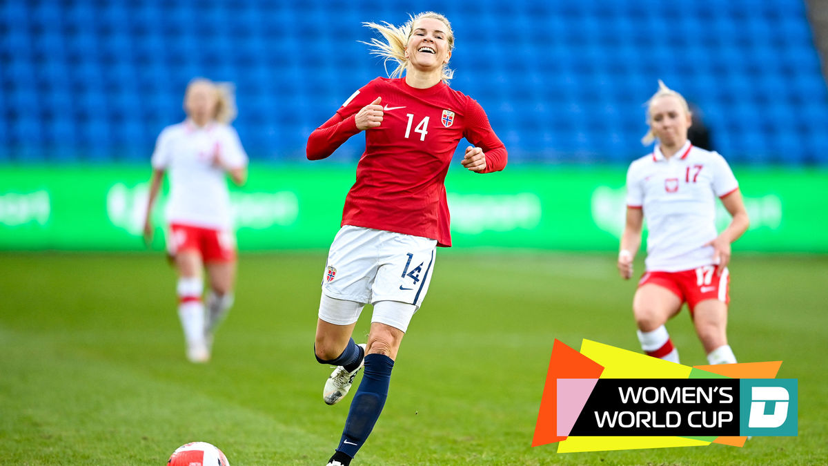 A white woman with a blonde ponytail and a red uniform top with white shorts smiles while dribbling a soccer ball past two women in white uniforms and red shorts.
