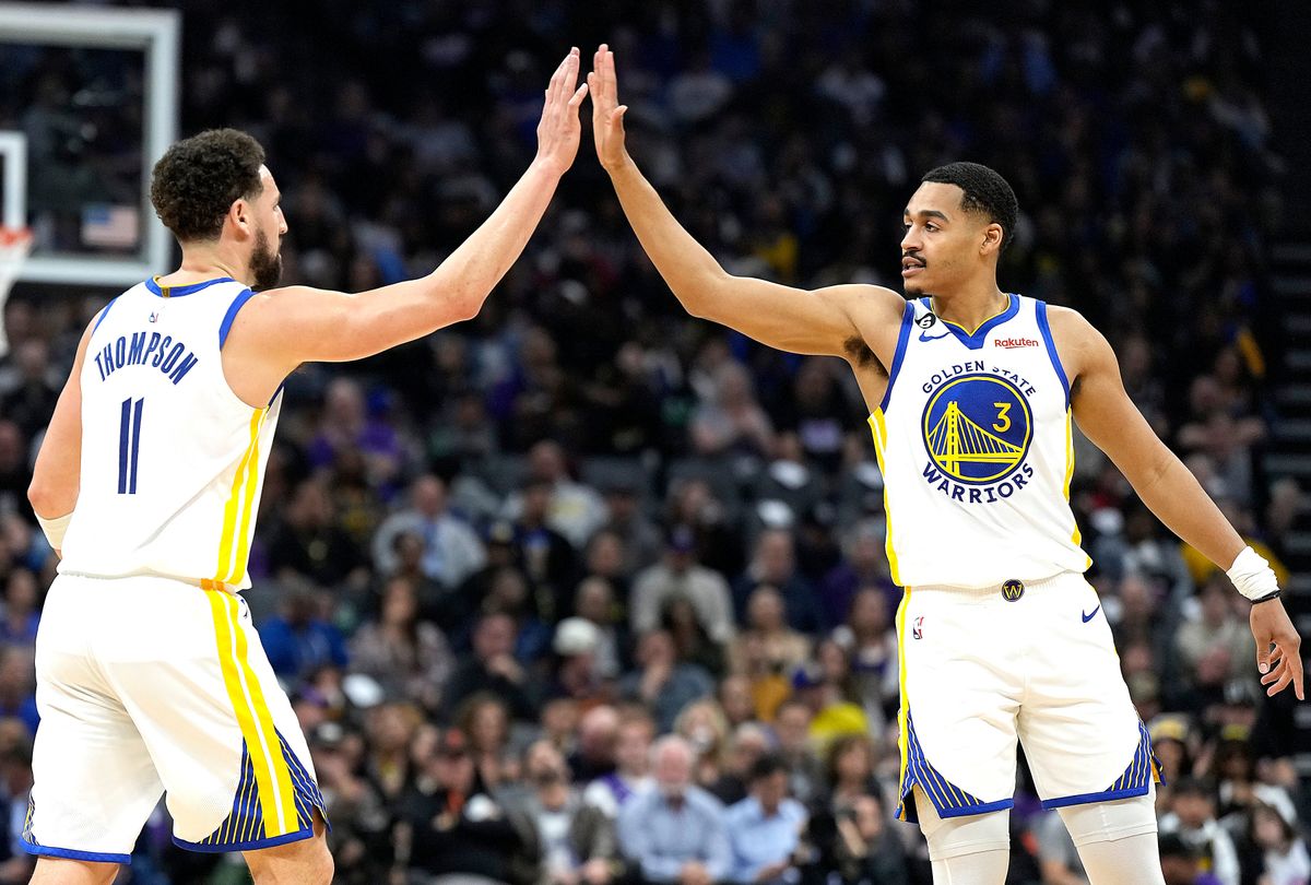 Two Black men in matching white uniforms with blue and white detailing, high five in the middle of an NBA basketball court while a crowd looks on.