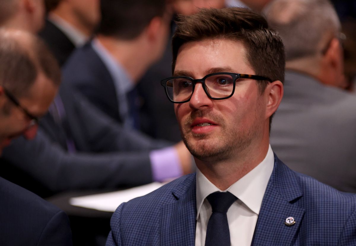 A white man in a blue suit and black tie, with stubble on his chin and prominent black glasses, sits with a bewildered look on his face.,