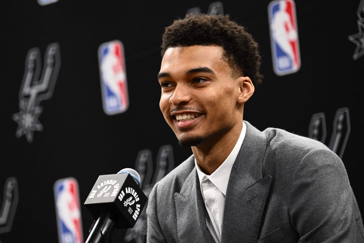 A young Black man in a gray suit in front of a black and white background smiles into a microphone at a San Antonio Spurs press conference.