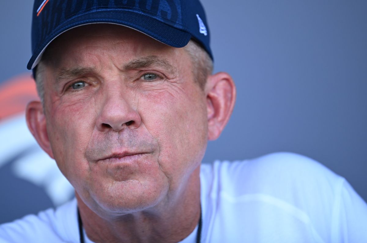 A white man in a navy blue hat and white t-shirt, with a whistle around his neck, purses his lips towards the camera.