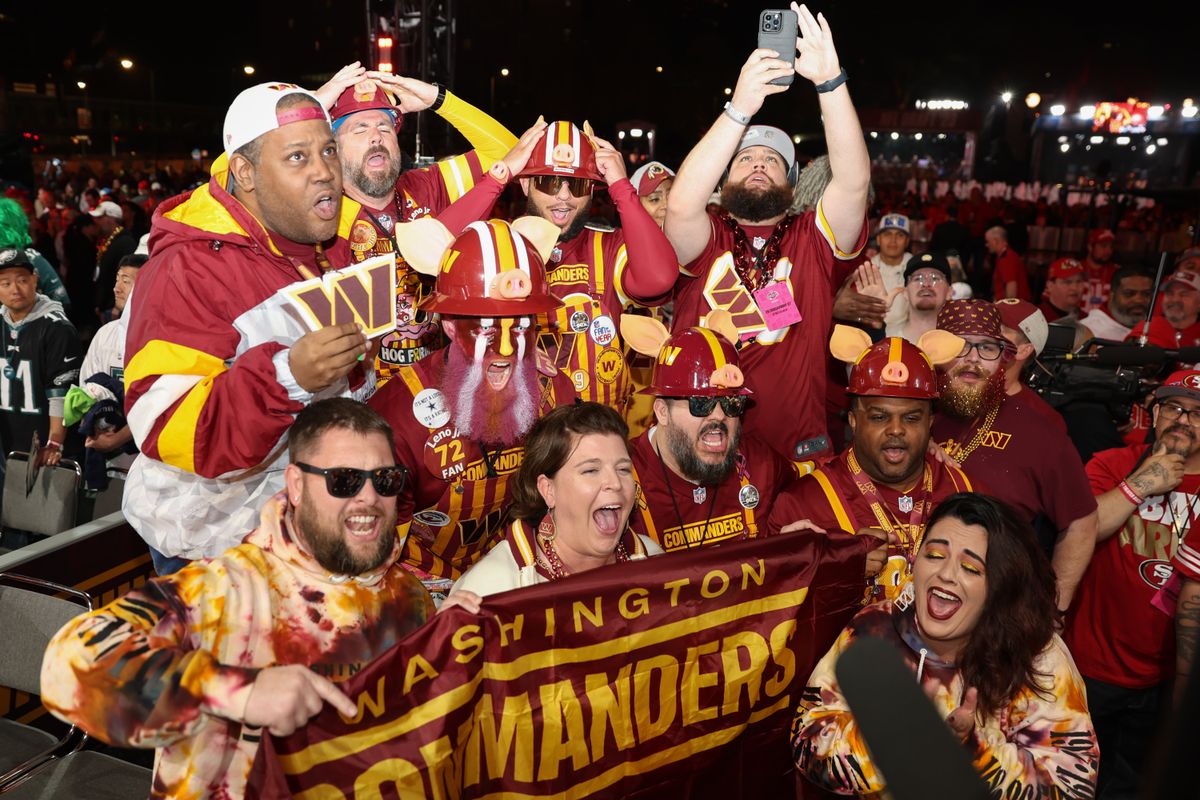 Several men and women, all dressed in maroon and gold, hold a Washington Commanders banner and yell at the NFL Draft.