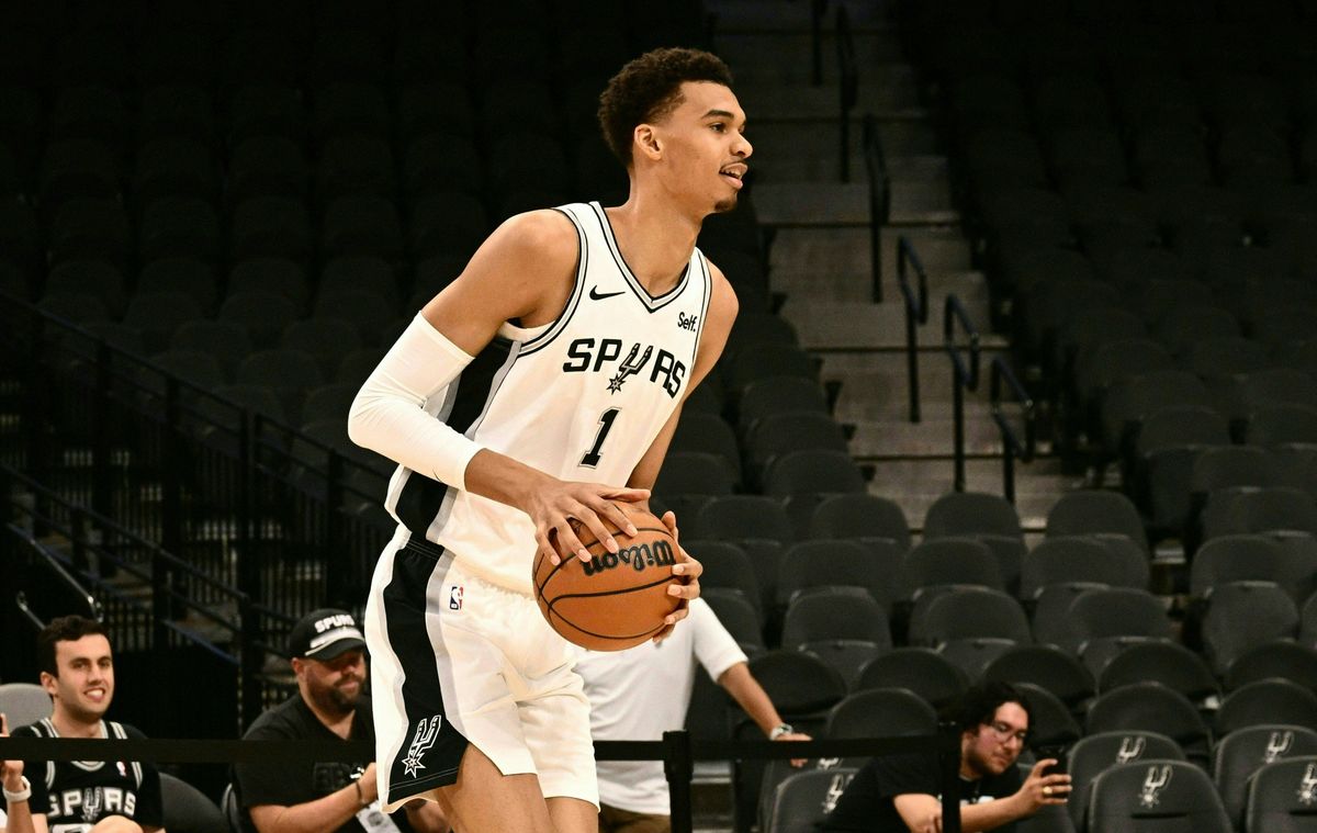 A tall, thin, young Black man wears a white San Antonio Spurs jersey and holds a basketball in an empty NBA arena with black seats. Three white men look on, one recording with his cell phone.