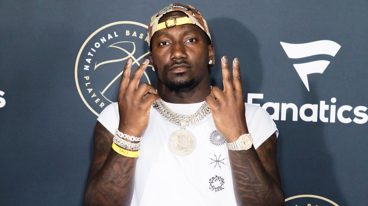 A Black man in a white t-shirt and backwards baseball cap holds up two fingers on each hand in front of a blue and white backdrop.