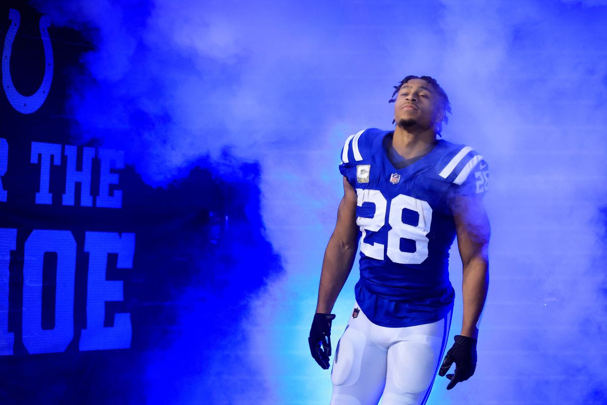 A black man with locs, wearing a blue Colts jersey and white pants, emerges through a cloud of blue smoke at an NFL football game.