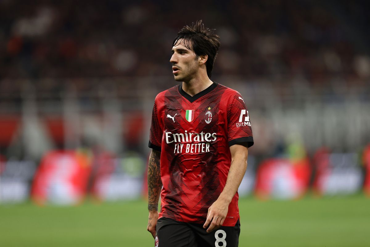 A white man with dark hair and a red and black jersey with black shorts and the number 8 walks on a soccer field.