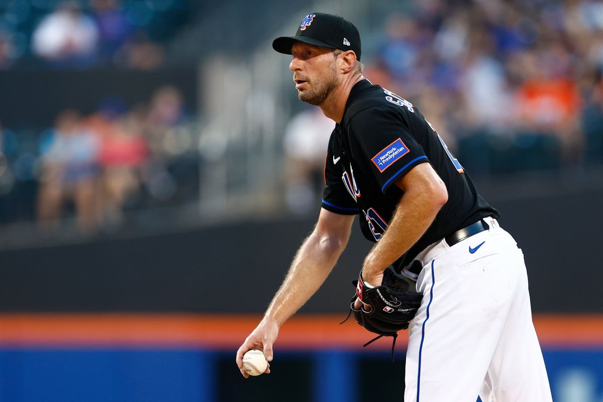 A white man in a black baseball jersey with white pants and a black hat holds the ball and looks towards the catcher on the pitchers' mound in a baseball stadium.