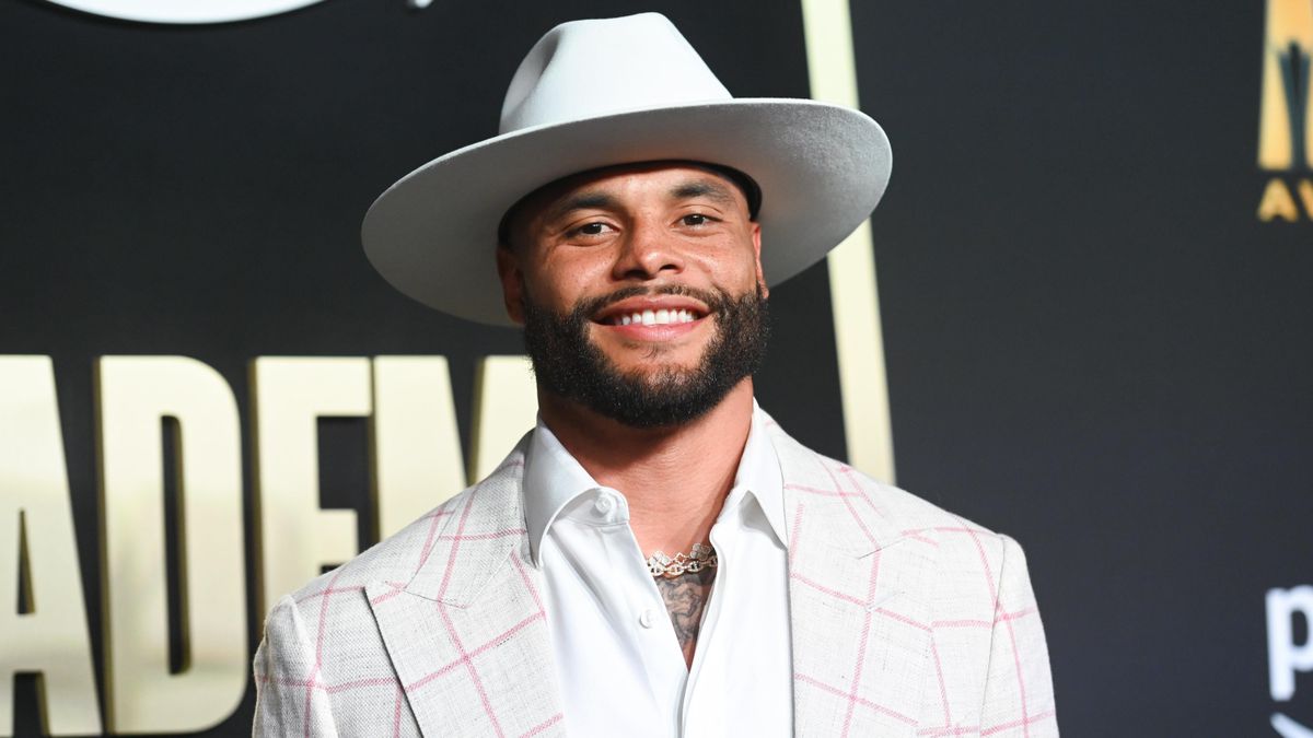 A Black man in a white broad-brimmed hat and pink and white suit coat with pink plaid smiles at the camera.