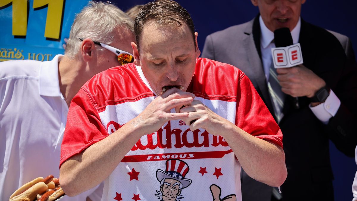 A white man with brown hair and a red and white shirt with a picture of Uncle Sam and "Nathan's" on it eats a hot dog with both hands.