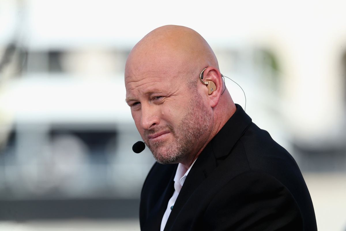 A bald white man with an ear piece and a microphone, wearing a black suit, squints while listening to a question on a broadcasting set.