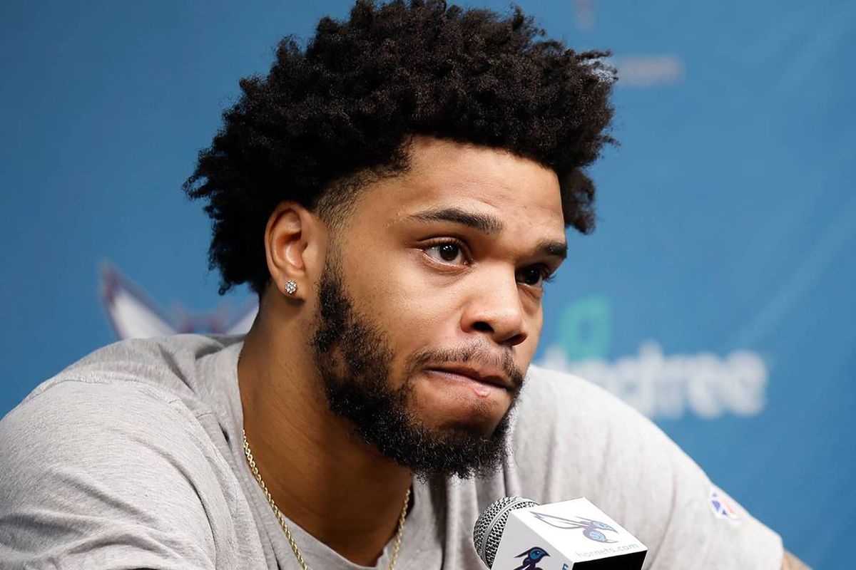 A young Black man with curly hair in a gray t-shirt bites his lip as he listens to a question before a microphone bearing a Charlotte Hornets logo.
